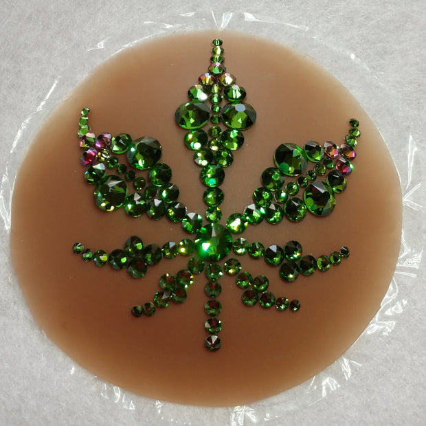 A luscious bright green marijuana leaf design made with various sizes of Fern Green Swarovski crystals & a few accents in a Green to Purple dynamic color shifting Swarovski crystal shade, on top of a dark flesh tone colored circular piece of concave silicone with a thin piece of plastic behind it. this object sits on a white textured surface