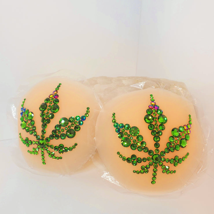 A luscious bright green marijuana leaf design made with various sizes of Fern Green Swarovski crystals & a few accents in a Green to Purple dynamic color shifting Swarovski crystal shade, on top of a light flesh tone colored circular piece of concave silicone with a thin piece of plastic behind it. this object sits on a white textured surface