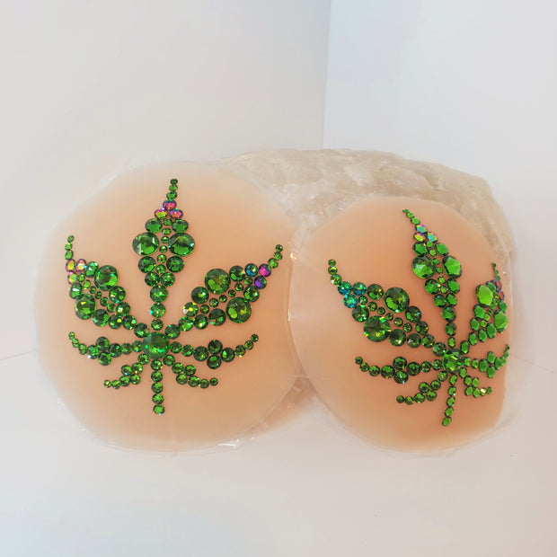 A luscious bright green marijuana leaf design made with various sizes of Fern Green Swarovski crystals & a few accents in a Green to Purple dynamic color shifting Swarovski crystal shade, on top of a medium-light flesh tone colored circular piece of concave silicone with a thin piece of plastic behind it. this object sits on a white textured surface